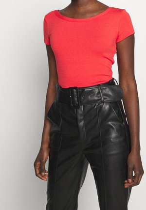 Žena ve vypasovaném korálově zbarveném tričku s krátkým rukávem zastrčeném do černých kožených kalhot s vysokým pasem a širokým páskem.