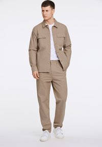Beige cotton jacket with two chest pockets and matching beige trousers. Model wearing a white t-shirt and white sneakers.