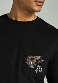 Schwarzes Langarmshirt mit einer strukturierten Tasche und einem gestickten Drachenmotiv in Orange, Schwarz und Weiß. Rundhalsausschnitt.