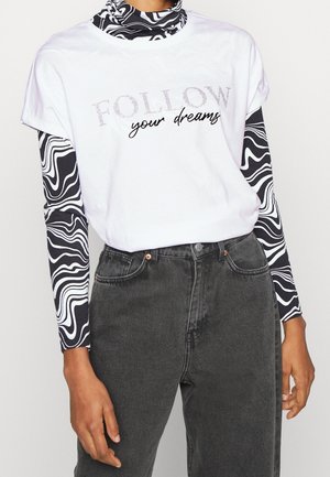 Weißes Baumwoll-T-Shirt mit dem Aufdruck "FOLLOW your dreams" in Grau und Schwarz, darüber getragen mit einem langärmeligen Oberteil im schwarz-weißen Wellenmuster, kombiniert mit dunklen Jeans.
