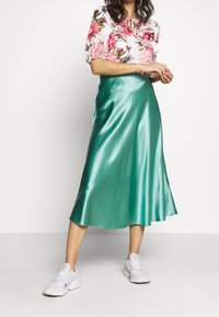 Mátově zelená saténová midi sukně s hladkou texturou, spárovaná s květinovou blůzou a bílými teniskami. Sukně má splývavý střih.