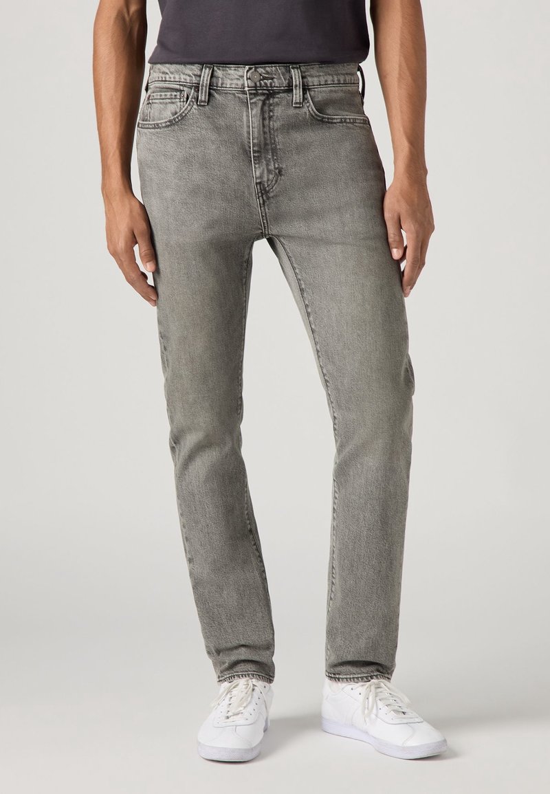 Homme portant un jean slim gris délavé, un t-shirt noir et des baskets blanches, debout devant un fond clair uni.
