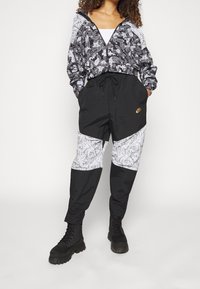 Černo-bílá vzorovaná zipová bunda s květinovým designem a odpovídajícími černými joggingovými kalhotami s paisley detaily; doplněno černými botami.