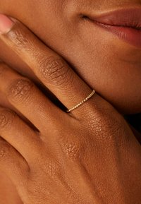 Bague en or avec une surface texturée, portée sur une main à la peau brunâtre, mettant en valeur une peau lisse et un vernis à ongles naturel.