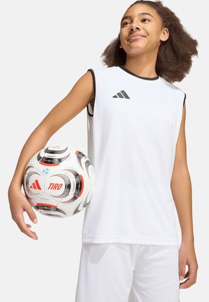 Młoda osoba w białym stroju sportowym trzyma białą piłkę nożną Adidas Tiro pod jednym ramieniem, patrząc lekko w górę z pewnym wyrazem twarzy.