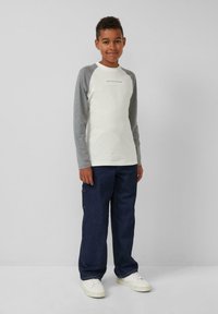 Weißes Langarmhemd mit grauen Raglanärmeln, kombiniert mit dunkelblauen Jeans und weißen Sneakers. Zu den Details gehört ein Logo auf dem Hemd.