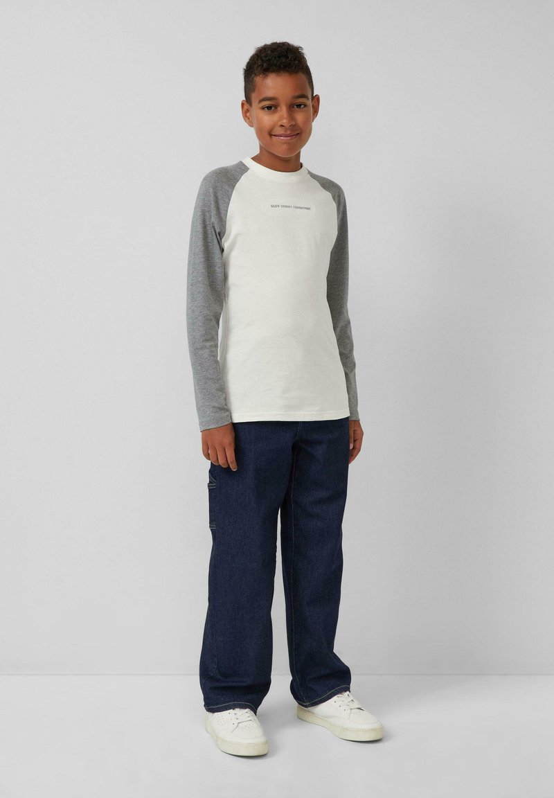 Weißes Langarmhemd mit grauen Raglanärmeln, kombiniert mit dunkelblauen Jeans und weißen Sneakers. Zu den Details gehört ein Logo auf dem Hemd.