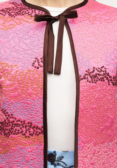 Ροζ μπουφάν με καπιτονέ σχέδιο και πολύχρωμη κεντητή διακόσμηση, καφέ περιθώριο και δέσιμο με φιόγκο πάνω από λευκή ριμπ μπλούζα και μπλε παντελόνι με σχέδιο.