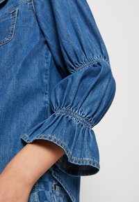 Denimtop med puffärmar, med en samlad elastisk nederkant. Tyget är blått, med en slät yta och kontrasterande sömdetaljer.