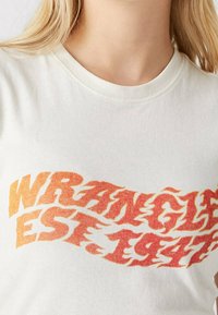 Bílý bavlněný tričko s vlnitým, přechodovým oranžovým grafickým vzorem, na kterém je tučným písmem napsáno "WRANGLER EST. 1947" s texturovaným povrchem.