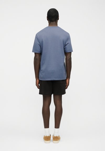 Άνδρας που στέκεται με την πλάτη προς τα εμπρός, φοράει μπλε μπλουζάκι, μαύρα σορτς, λευκές κάλτσες και καφέ αθλητικά παπούτσια, μπροστά σε λευκό, απλό φόντο.