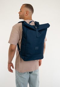 Zaino navy roll-top realizzato in tessuto resistente, dotato di spallacci regolabili, tasca frontale con zip e una toppa con logo sul pannello frontale.
