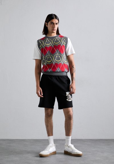 Gestrickte graue Weste mit rot-gelbem Rautenmuster, kombiniert mit schwarzen Shorts mit einem Bärenmotiv und weißen Turnschuhen mit beiger Sohle.
