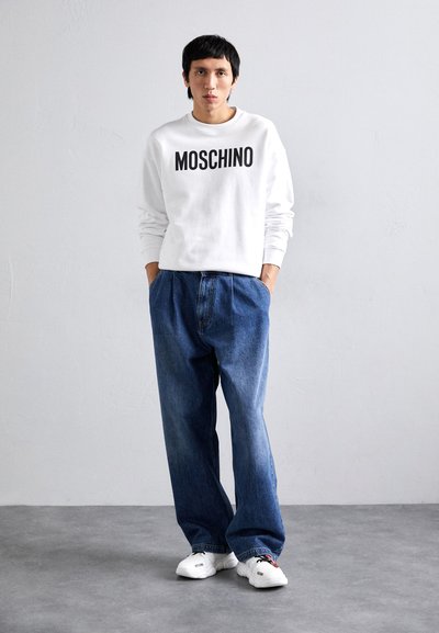 Hvit genser med svart "MOSCHINO" tekst, kombinert med løse blå denimbukser og hvite sneakers. Casual, avslappet passform.