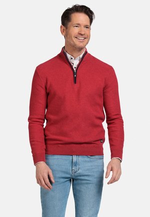 Rode gebreide trui met een halve rits kraag, contrasterende marineblauwe afwerking, geribbelde manchetten en zoom. Draagt met lichtblauwe spijkerbroek en een gemusterd shirt eronder.