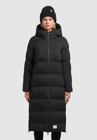 Cappotto invernale imbottito nero con una silhouette lunga, chiusura con zip, cappuccio e tasche laterali. Berretto nero lavorato a maglia con un piccolo dettaglio del logo.
