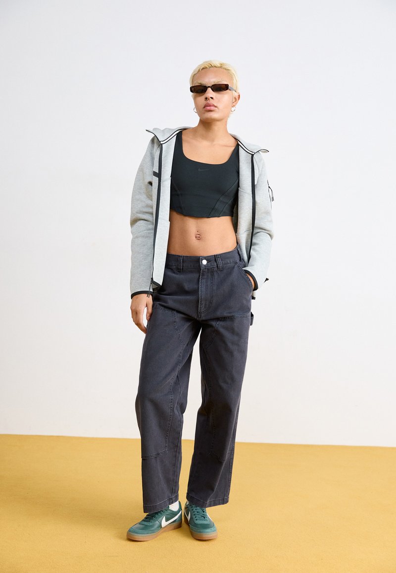 Sudadera gris con cremallera, top corto negro, pantalones de pierna ancha oscuros, zapatillas verdes con acentos blancos y gafas de sol oscuras. Tejido suave y texturas distintas.