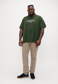 Alyvuogių spalvos per didelis marškinėliai su balta "LEVI'S" logotipu, derinami su šviesiai smėlinėmis kelnėmis ir juodais sportbačiais su guminiu padu.
