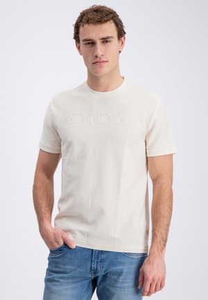 Młody mężczyzna z krótkimi kręconymi włosami nosi kremowy t-shirt z krótkim rękawem z subtelnym logo i niebieskie dżinsy, stoi na jednolitym tle.