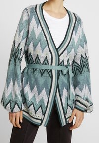 Stickad kofta med tealfärgad, vit och svart zigzagmönster, ballongärmar och ett matchande bälte, bärs över en vit skjorta och mörka byxor.