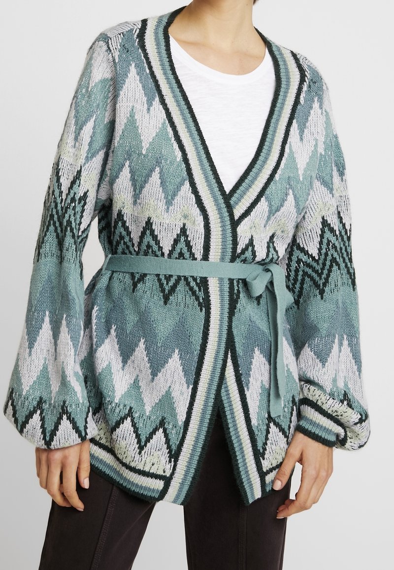 Stickad kofta med tealfärgad, vit och svart zigzagmönster, ballongärmar och ett matchande bälte, bärs över en vit skjorta och mörka byxor.