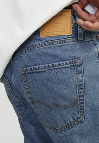 Blå denim-bukser med et læderlap på taljen, med orange syninger og baglommer med buet detaljering.