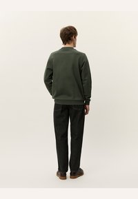 Sudadera verde oscuro con cuello redondo y mangas largas, vista desde la parte de atrás, combinada con pantalones negros casuales y zapatos marrones.