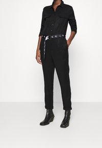 Combinaison noire à manches longues, avec deux poches poitrine et une ceinture à motifs. Design ajusté avec des jambes fuselées et des bottes à talons noires.