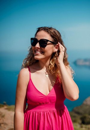 Růžové bezrukávové šaty s upnutým pasem, doplněné velkými černými slunečními brýlemi a náhrdelníkem s přívěskem; kudrnaté vlasy. Pozadí oceánu.