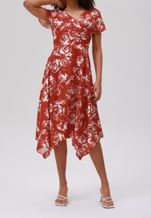 Vrouw draagt een knielange jurk met roestoranje en witte bloemenprint, korte mouwen en een puntige zoom, gecombineerd met witte sandaaltjes met bandjes.