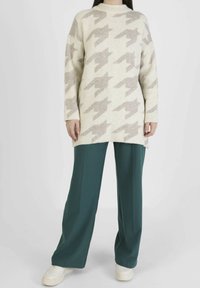 Personne portant un pull crème à motif pied-de-poule gris clair, un pantalon large vert foncé et des baskets blanches sur un fond blanc.