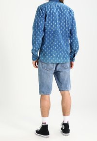 Blauwe spijkerbutton-up shirt met wit bloemenpatroon, gedragen met lichtblauwe spijker shorts en zwarte hoge sneakers, gezien vanachter.