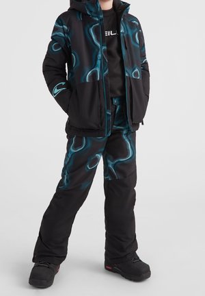 Barn iført sort og blå mønstret vinterjakke og bukser med sorte støvler, stående med hænderne i lommerne på grå baggrund.