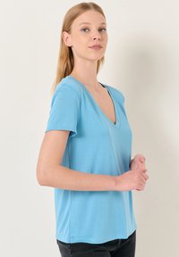 Błękitna koszulka z krótkim rękawem z dekoltem w kształcie litery V, wykonana z gładkiego materiału, o luźnym kroju i subtelnych detalach przeszycia.
