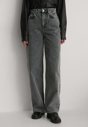 Osoba nosząca szare dżinsy z wysokim stanem i luźnym krojem, zestawione z czarnymi masywnymi loafersami oraz czarną bluzką z długim rękawem na jednolitym tle.