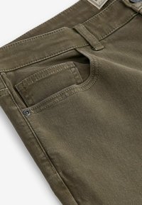 Gros plan sur un jean vert olive montrant la poche avant, la passoire de ceinture, le rivet et les coutures détaillées sur le tissu en coton.