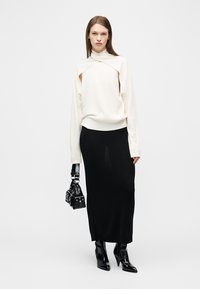Γυναίκα με λευκό πουλόβερ με κοψίματα, μακριά μαύρη φούστα, μαύρα μπότες με τακούνι, κρατώντας μαύρη τσάντα με τρούκς, στέκεται μπροστά σε λευκό φόντο.