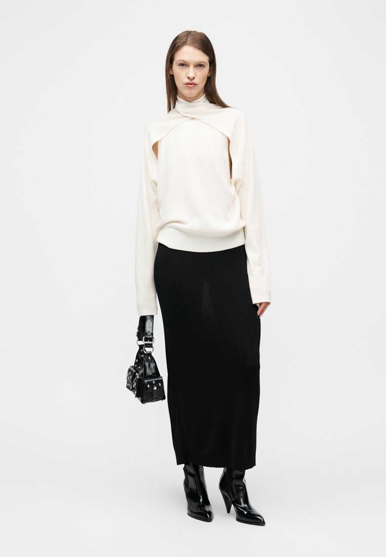 Femme portant un pull blanc à découpe, une longue jupe noire, des bottines noires à talons, tenant un sac à main noir clouté, debout devant un fond blanc.