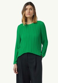 Blouse plissée verte à manches longues, col rond et coupe décontractée. Texture lisse avec des rayures verticales, associée à un pantalon sombre.