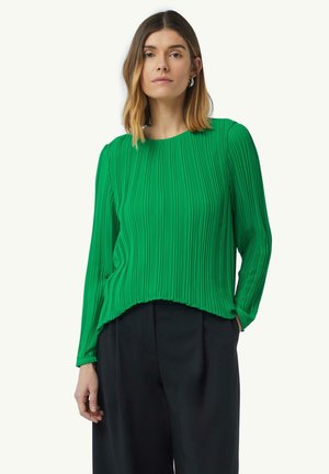 Blouse plissée verte à manches longues, col rond et coupe décontractée. Texture lisse avec des rayures verticales, associée à un pantalon sombre.