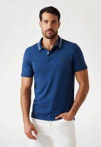 Uomo con capelli corti e scuri, che indossa una camicia polo blu e pantaloni bianchi, in piedi con una mano in tasca su uno sfondo semplice.