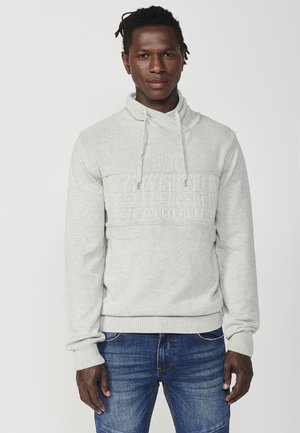 Lichtgrijze sweatshirt met een funnelnek, textuurstof en een patroonaccent op midden van de borst. Gecombineerd met blauwe jeans.
