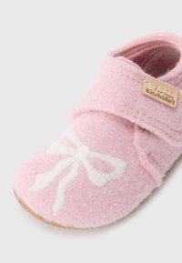 Rožinė veltinio kūdikių batukas su baltu siuvinėtu kaspinėliu, Velcro dirželiu ir rudais padukais. Pasižymi minkšta tekstūra ir lanksčiu dizainu.