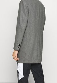 Grijze jas van een wolmix met een getailleerde pasvorm, zwart gekleurde knopen aan de manchetten en een rechte snit, weergegeven van achteren.