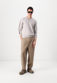 Pull gris avec des rayures contrastantes rouges et bleu marine sur les manches, pantalon beige plissé, et bottes marron à lacets. Des lunettes de soleil complètent le look.