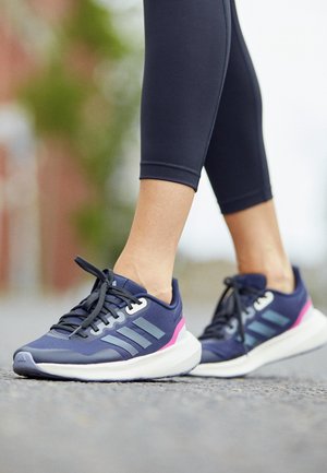 Pies con leggings negros que llevan zapatillas azul marino con rayas azul claro y detalles rosas, de pie sobre una superficie pavimentada al aire libre.