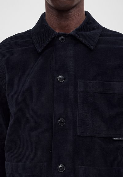 Homme portant une veste-chemise en velours côtelé bleu marine foncé avec des boutons noirs et une grande poche avant sur le côté gauche.