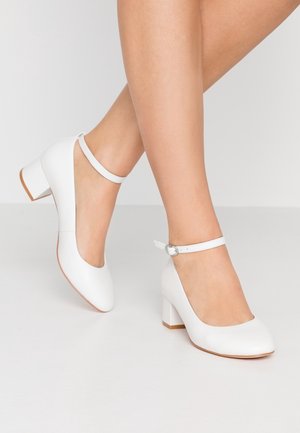 Białe skórzane buty na słupku z zaokrąglonym noskiem i paskiem wokół kostki. Gładka faktura z delikatnym połyskiem i kontrastującą brązową podeszwą.