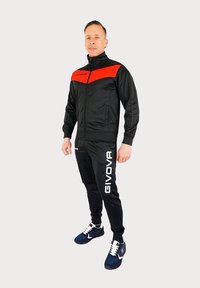 Zwarte trainingsoutfit met een rood chevronontwerp op de jas. Voorzien van GIVOVA-branding in het wit. Gepaarde met marineblauwe sneakers.