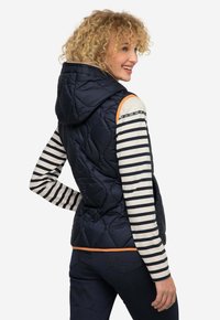 Gilet blu navy imbottito con cappuccio, caratterizzato da dettagli arancioni e zip. Indossato sopra una maglietta a righe a maniche lunghe e abbinato a jeans scuri.
